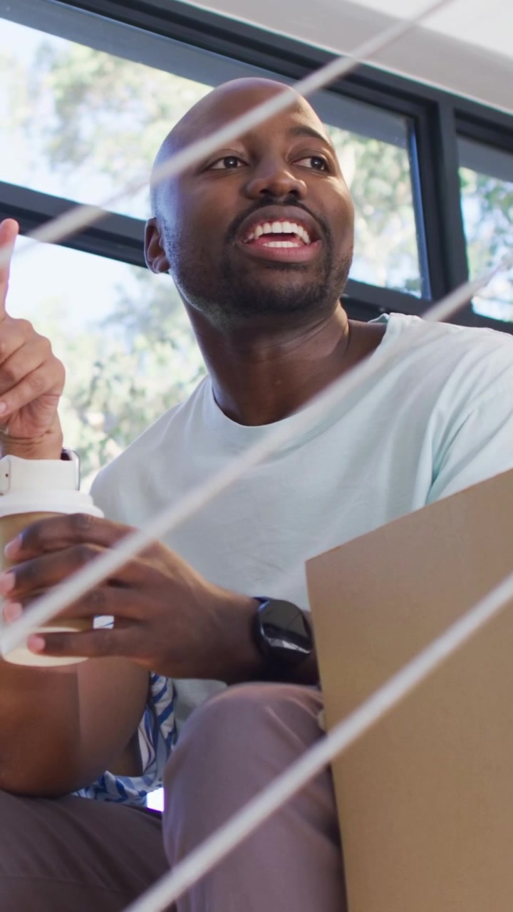 Vertical video: Smiling man wearing tee on stairs offering cup after friend prompts, reaching box
