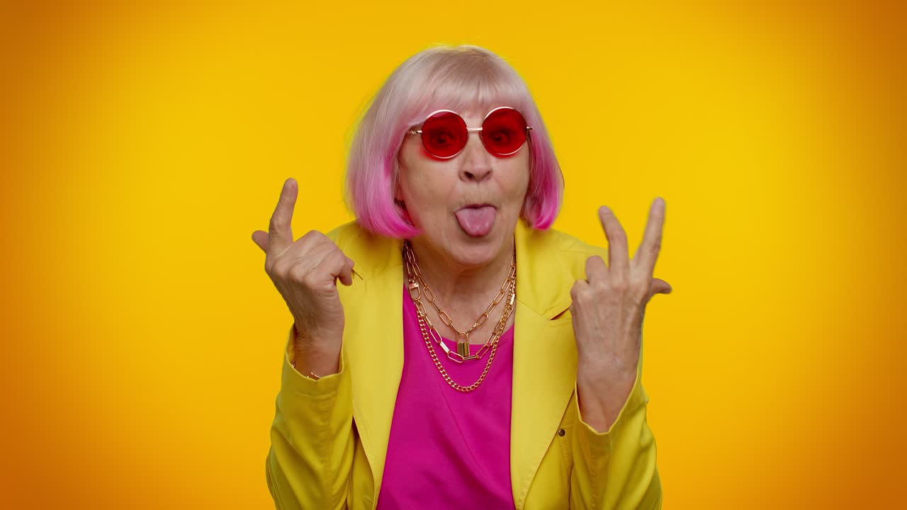 una mujer mayor con cabello rosa, blazer amarillo y gafas de sol rosas sonríe y señala con sus dedos índice