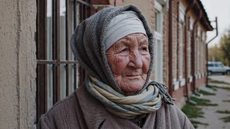 weathered grandmother wearing traditional headscarf standing on quiet village street, gazing pensively toward distant horizon with deep, reflective wisdom