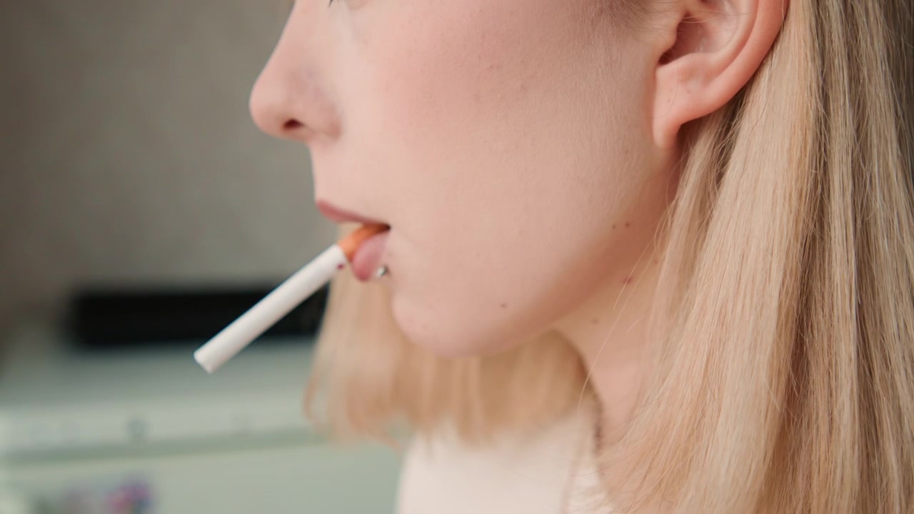 Vrouw met sigaret pauzeert vredig. Kalme Kaukasische vrouw met tatoeages ontspant bij zacht licht. Rustige vrouw met subtiele make-up en piercings houdt sigaret vast in zachte keukenambiance.
