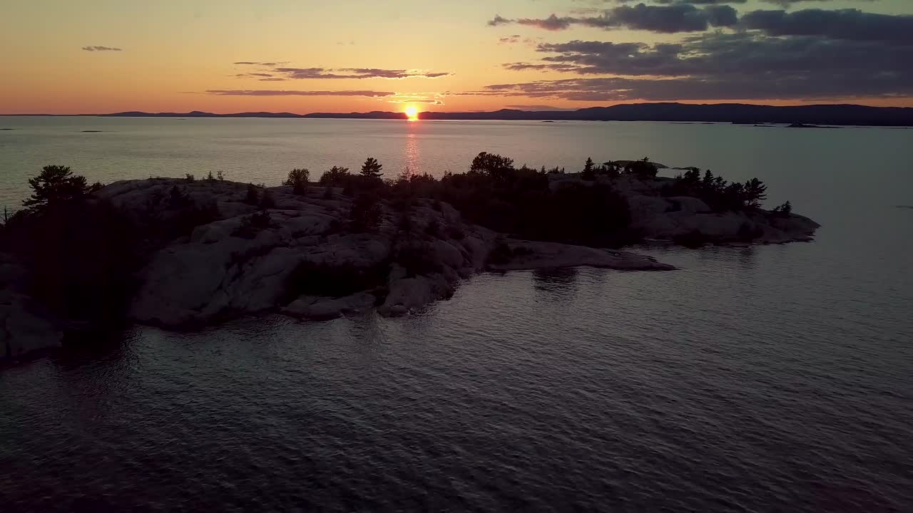Revelação de um pôr do sol épico sobre uma ilha rochosa com pinheiros em um lago azul, tomada aérea ampla de drone com dolly out.