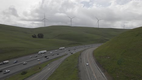 Bajo-Un-Cielo-Cubierto-De-Nubes,-Este-Metraje-De-Drones-Revela-El-Viaje-Tranquilo-De-La-Autopista-580-A-Través-De-Las-Colinas-Verdes-Salpicadas-De-Molinos-De-Viento-Del-Paso-De-Altamont,-Ligeramente-Salpicado-De-Tráfico-Escaso-Pero-Constante