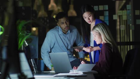 Video-of-three-diverse-male-and-female-colleagues-talking-around-a-laptop-at-night-in-office