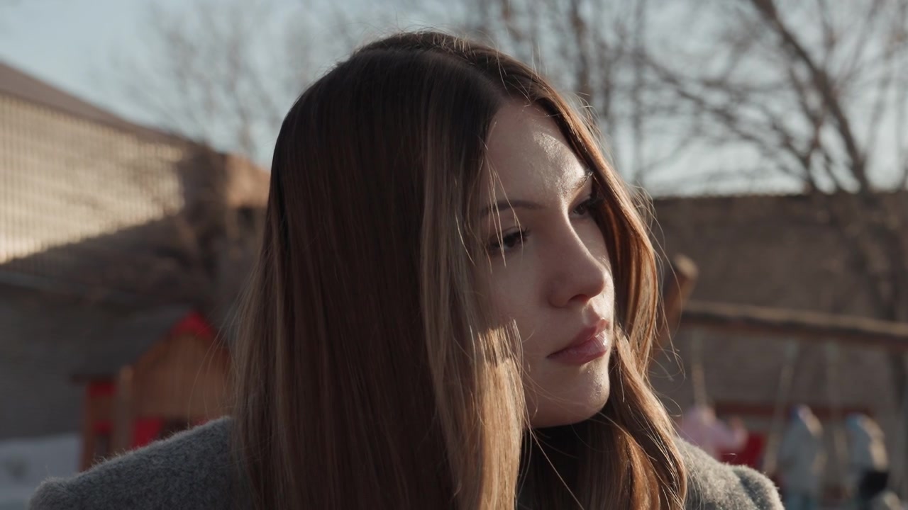 Ritratto sereno di bella donna alla luce del sole, occhi chiusi ed espressione dolce, capelli lunghi che catturano la luce di contorno, cappotto di lana e sfondo di parco giochi urbano dai colori tenui, texture della pelle delicata, stato d'animo calmo e riflessivo.