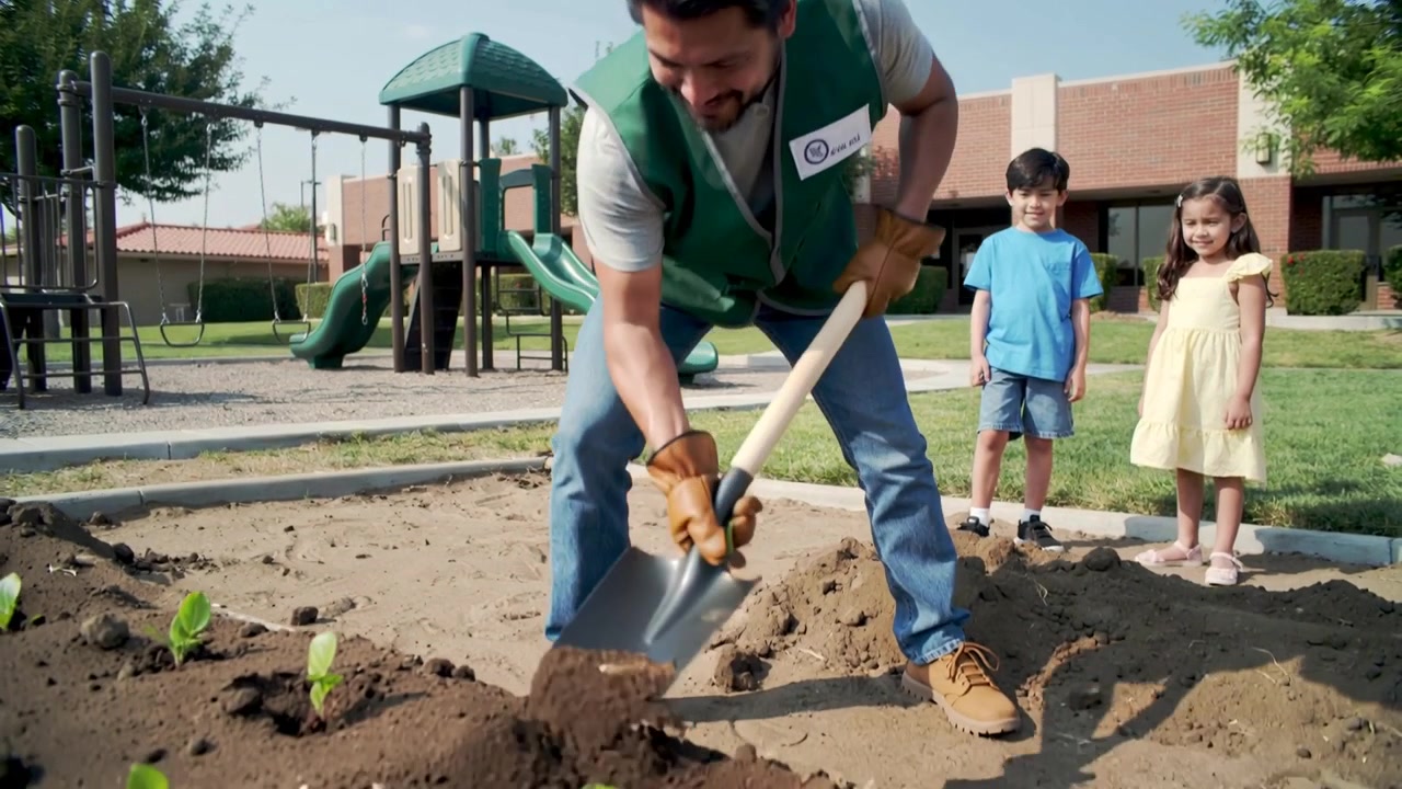 Leaning gloved worker in green vest placing plants at playground bed with shovel and kids watching
