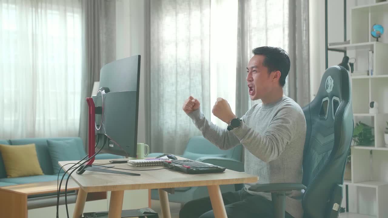 Asian Man In Long Sleeved T-Shirt And Black Pants Looking At Desktop Computer Screen And Being Happy After Winning The Project While Working At Home.