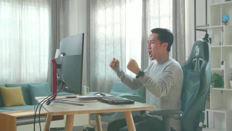 asian man in long sleeved t-shirt and black pants looking at desktop computer screen and being happy after winning the project while working at home.