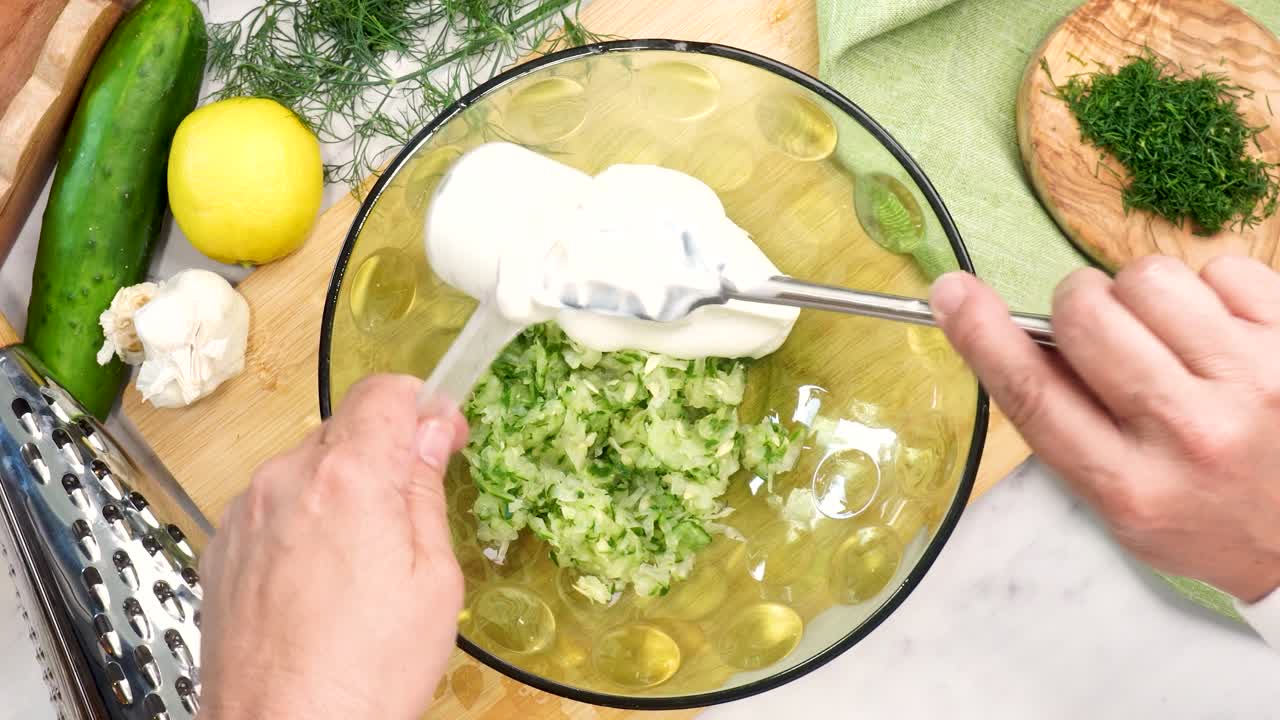 Making Tzatziki Greek or Mediterranean sauce or dip for a party. Dip or sauce in a green bowl on a wooden cutting board with dill, green napkin and a grater. Adding Greek yogurt to the Tzatziki sauce.