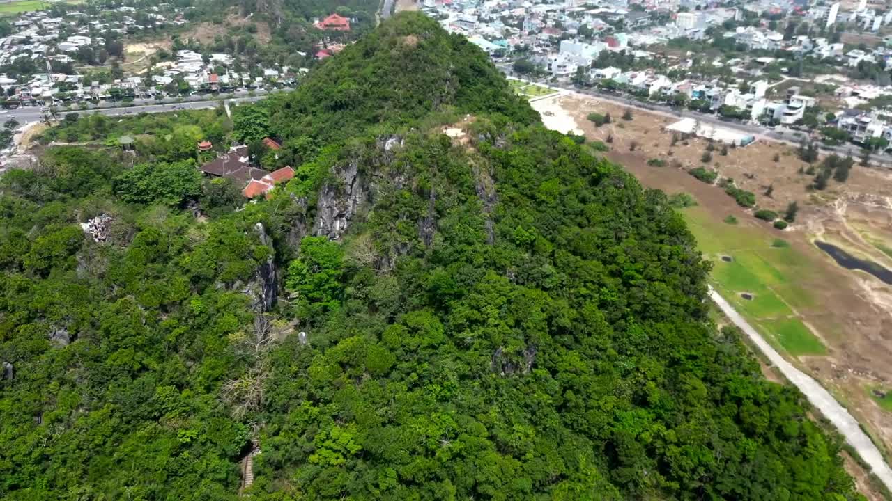Aerial view of green hill with Buddhist temples, red-tiled roofs, and surrounding city buildings in Vietnam, showcasing contrast between nature, tradition, and modern development.