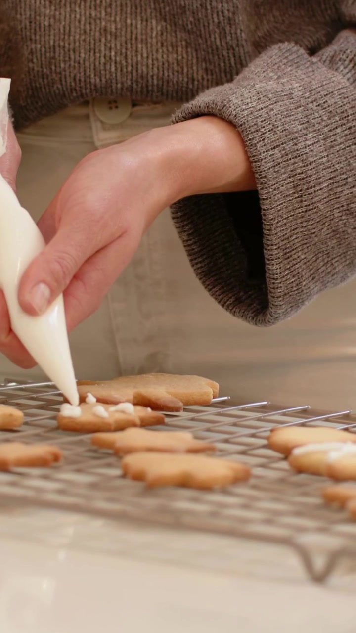 Video verticale: Donna sorridente che decora con precisione dei biscotti con una sac à poche a casa, indossando un maglione.