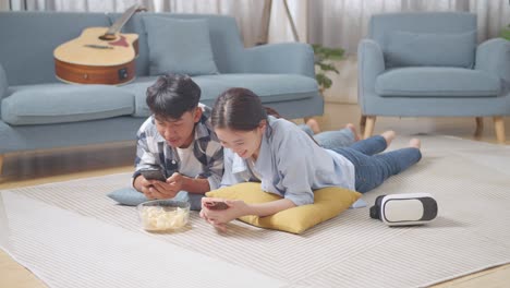 full body of asian teen couple playing smartphone while lying on carpet on the floor at home. phone addicted couple