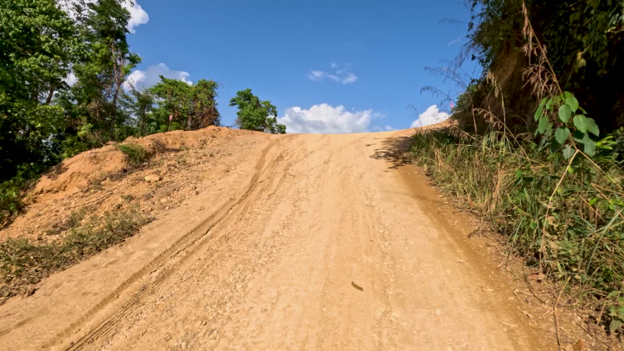 Vehicle travels up winding dirt road, surrounded by lush greenery, under bright midday sunlight