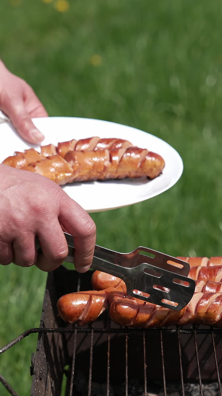 Chef taking off ready sausages from a grill. Man putting delicious roasted hot dog sausages on a plate with a tongs. Barbecue for outdoor lunch. Vertical video