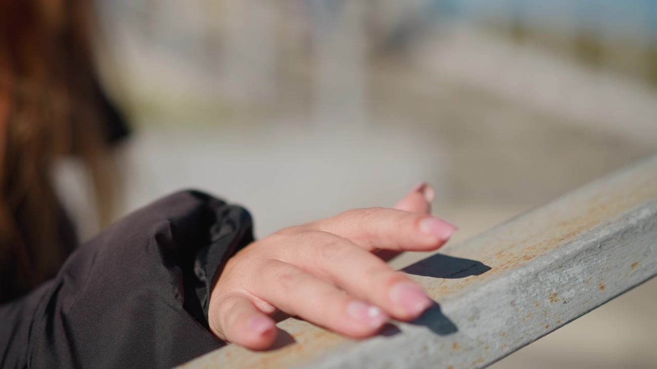 Mano di donna poggia su una ringhiera metallica invecchiata con una morbida luce naturale che evidenzia la texture della sua pelle e delle unghie dipinte. Lo sfondo rimane leggermente sfocato, enfatizzando il primo piano