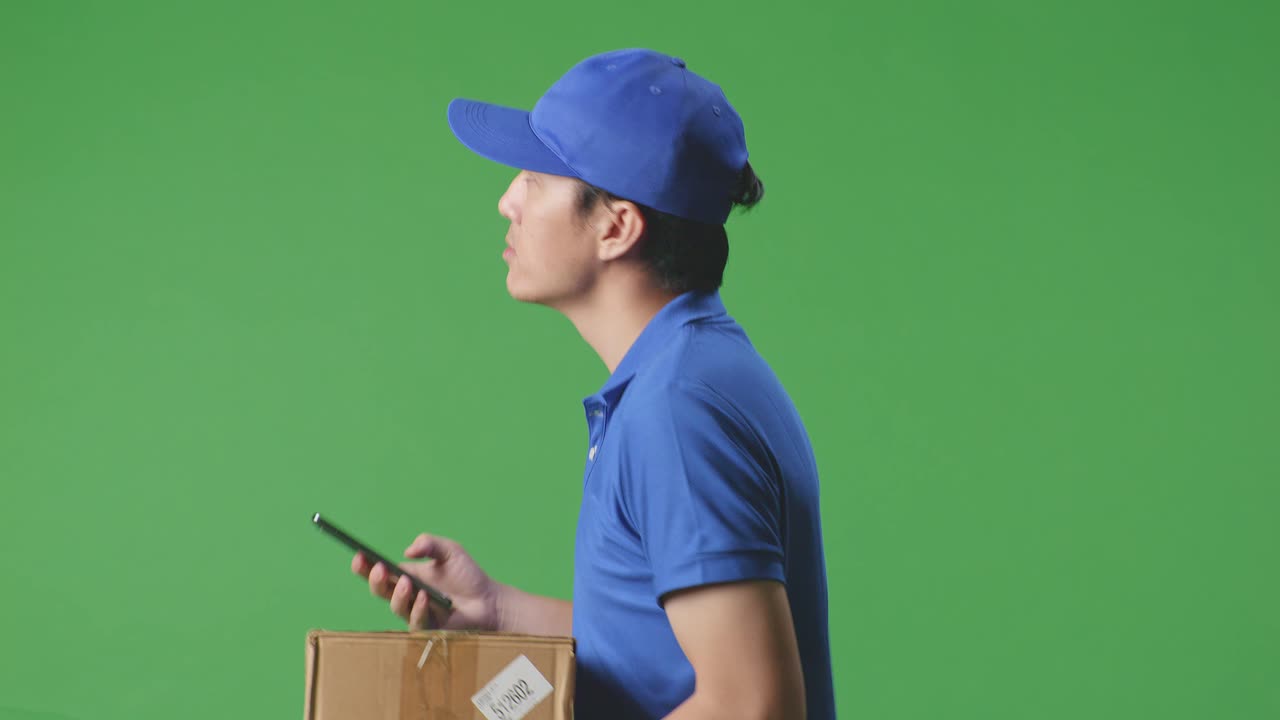 Close Up Side View Of Asian Male Courier In Blue Uniform Walking And Using Smartphone While Delivering A Carton On Green Screen Background In The Studio