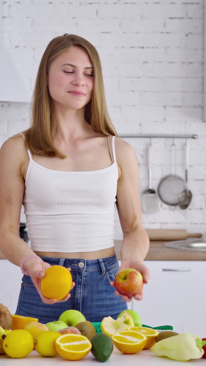 Portrait of beautiful girl in the kitchen. Vegetarian woman holding organic fruit. Happy young housewife near the table with fresh fruit and vegetables. Vertical video
