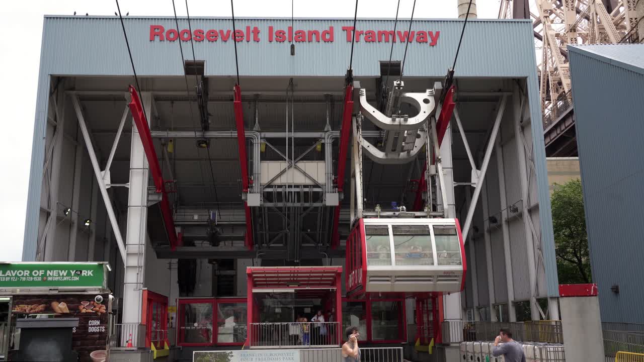 Steady medium shot of the Roosevelt Island Tramway cabin departing from the island station, with people walking in front as the cabin moves overhead