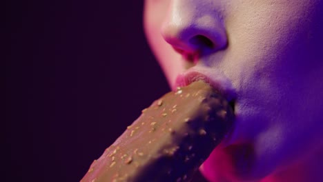 cerca mientras una joven bonita toma un bocado de un helado de chocolate vegano y disfruta del sabor mientras sonríe con contraste púrpura en su cara