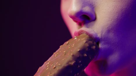 close up while a pretty young woman takes a bite of a vegan chocolate ice cream and enjoys the taste while smiling with purple contrast on her face