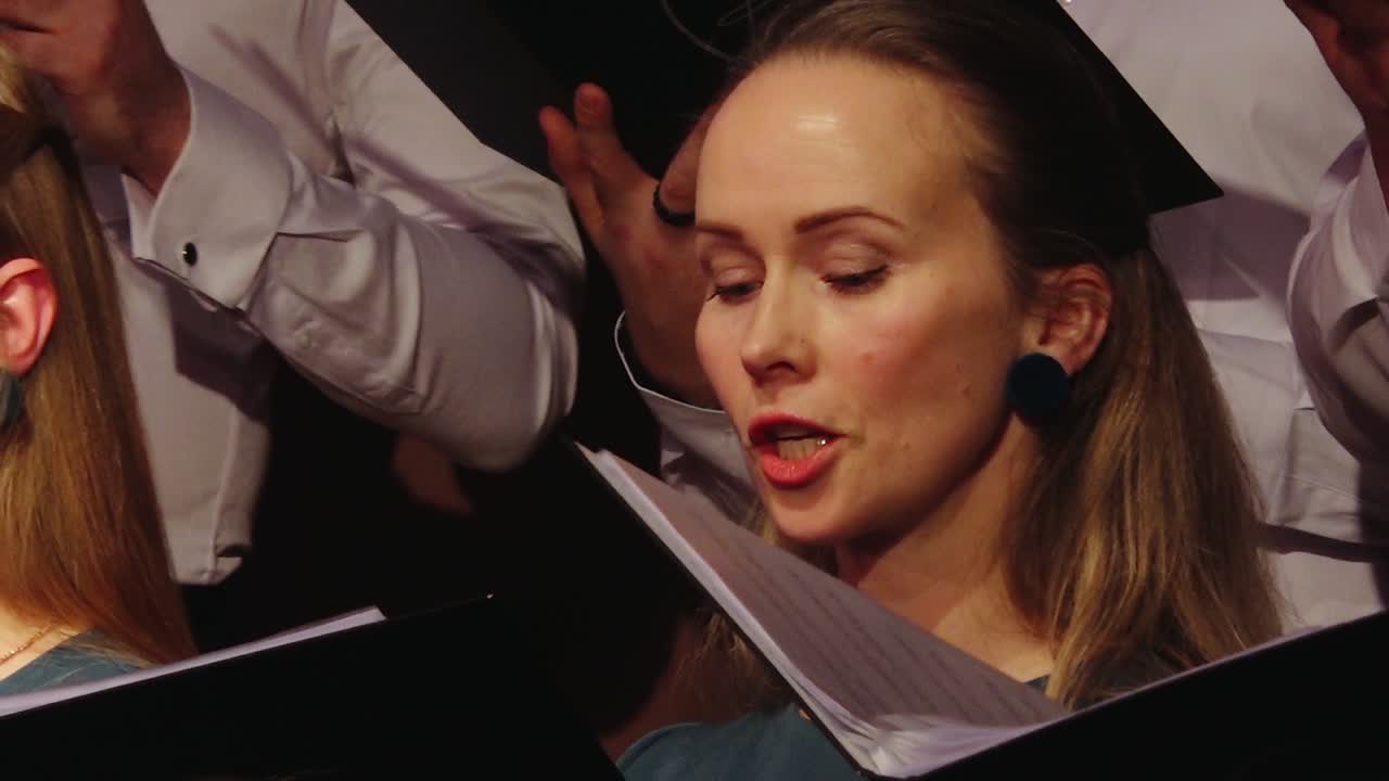A focused woman sings in a choir, holding sheet music as others around her perform, capturing a moment of vocal expression and concentration.