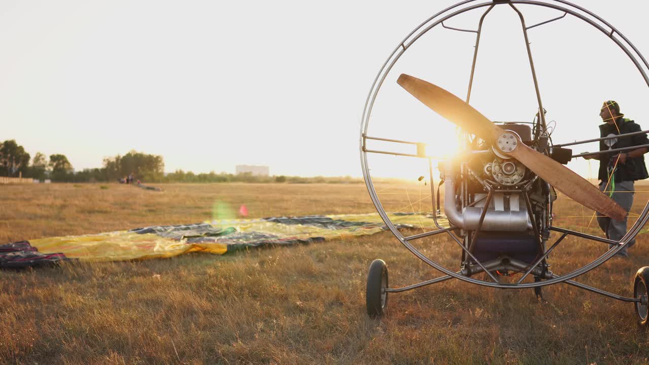 моторный парапланер стоит в поле при заходе солнца с деревянным пропелером, а пилот выкладывает парашют и выстраивает шланги