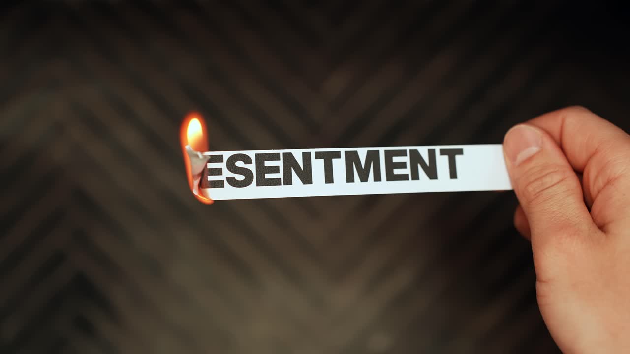 Em close-up, uma mão segura uma tira de papel com a palavra “RESSENTIMENTO” enquanto a borda se incendeia, simbolizando amargura e sentimentos não resolvidos em um fundo escuro com espaço para texto