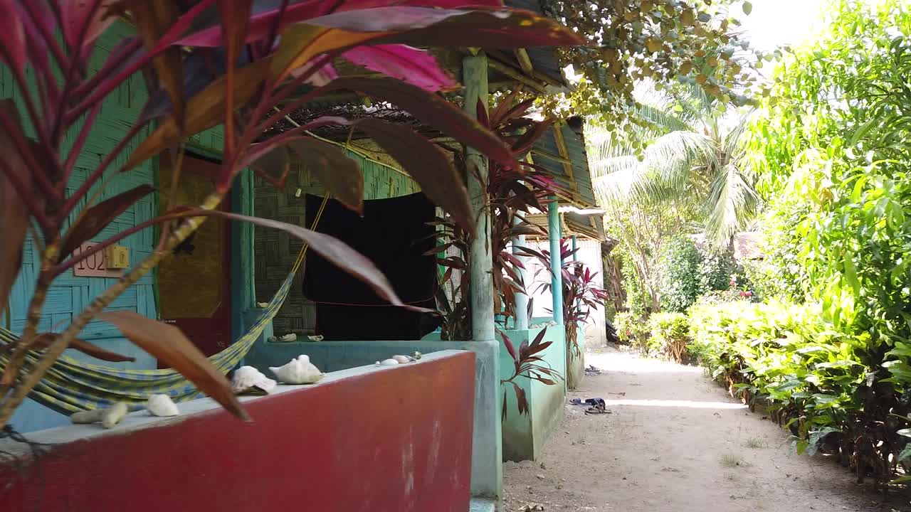 Slow motion swing from a guest house sleeping hut for rent with a hammock concrete wall reed panels and corrugated iron roof to the row of huts and the well maintained green garden around