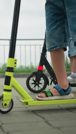 leg view of a child riding a green scooter beside their mother, who is carrying the younger child on a scooter