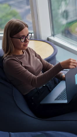 thoughtful young lady sitting near big window opens her laptop and starts work. woman typing on computer looking into window. high angle view. vertical video