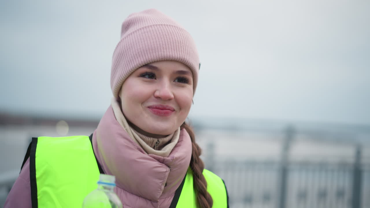 Young female worker wearing pink beanie, scarf, and reflective vest holding sandwich and water bottle outdoors on cold overcast day, looking ahead with thoughtful expression