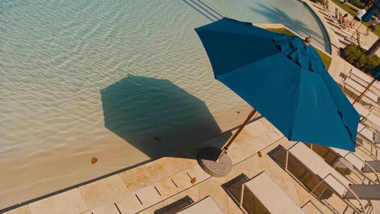 Panoramica del drone che mostra un ombrellone blu che proietta ombra su una piscina di un resort, ospiti in costume, spazio per il testo