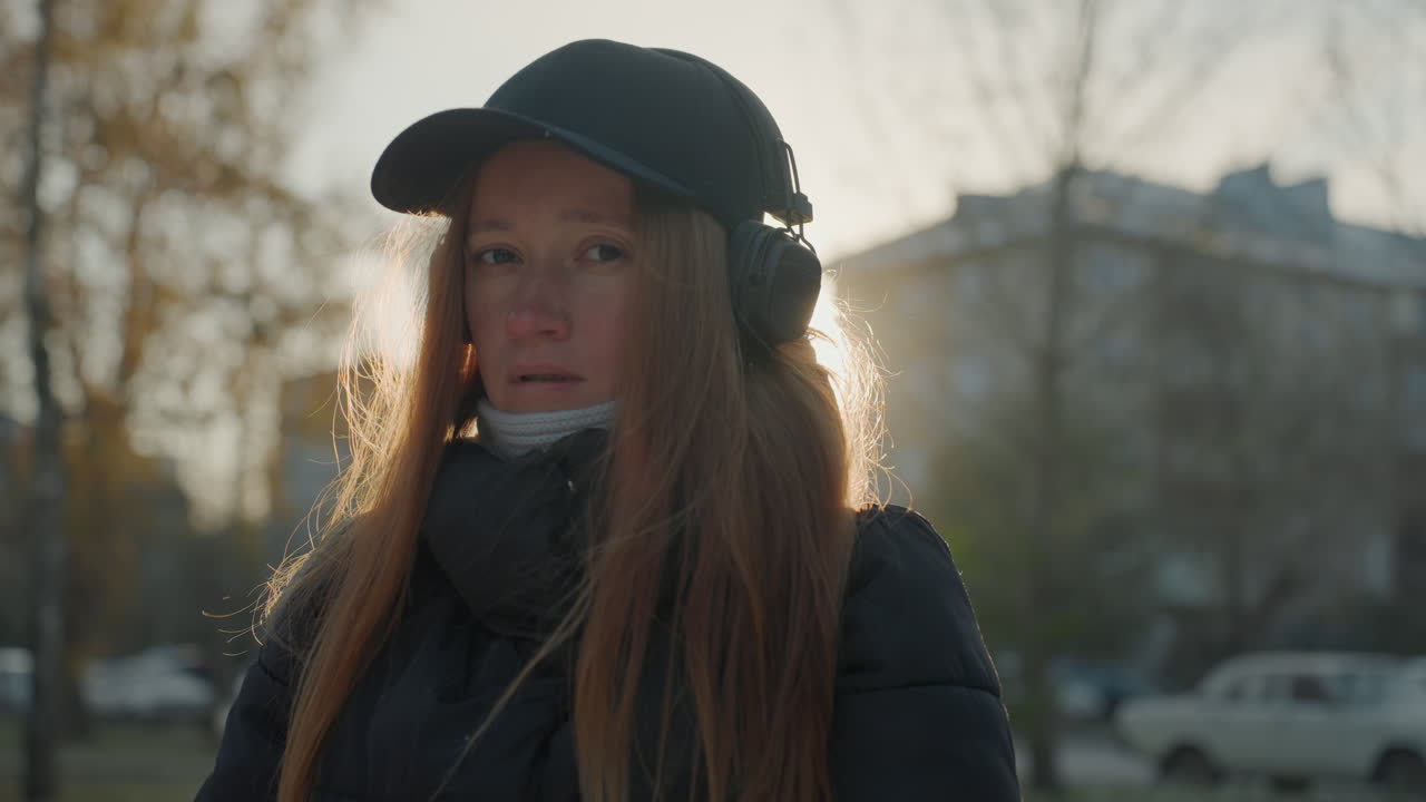 Ampia inquadratura di donna bianca in città. Pendolare che si aggiusta il guanto e ascolta con le cuffie, controluce dorato, auto ed edifici sullo sfondo, respiro visibile, sguardo cauto, umore contemplativo.