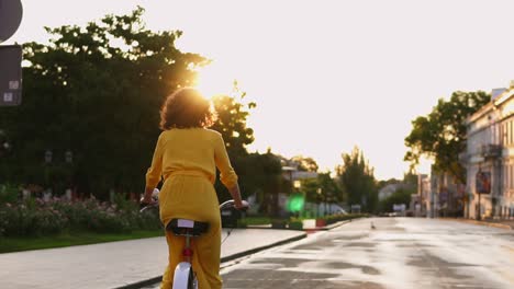 Vista-Posterior-De-Una-Mujer-Irreconocible-Con-Un-Largo-Vestido-Amarillo-Montando-Una-Bicicleta-En-El-Centro-De-La-Ciudad-Durante-El-Amanecer-Disfrutando-De-Ella