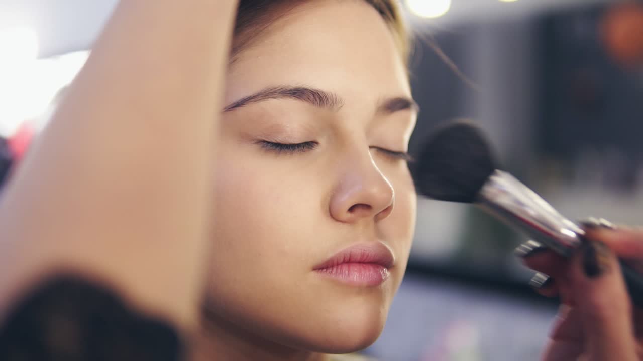 Professional make-up artist at work: Close Up view of female hands applying facial powder on young woman's skin using special