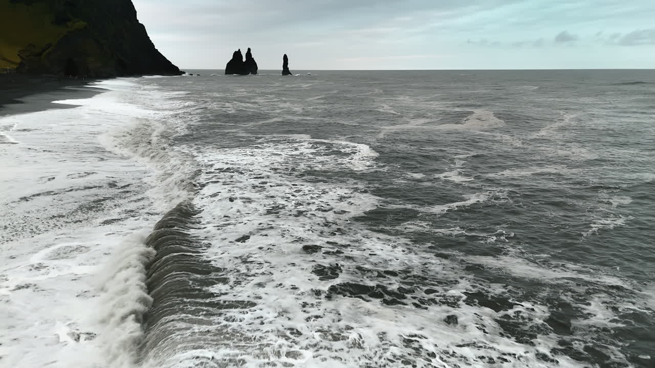 Wide foamy white waves arrive to the beach with black volcanic sand. Drone flight above the raging waterscape of the North Atlantic Ocean.