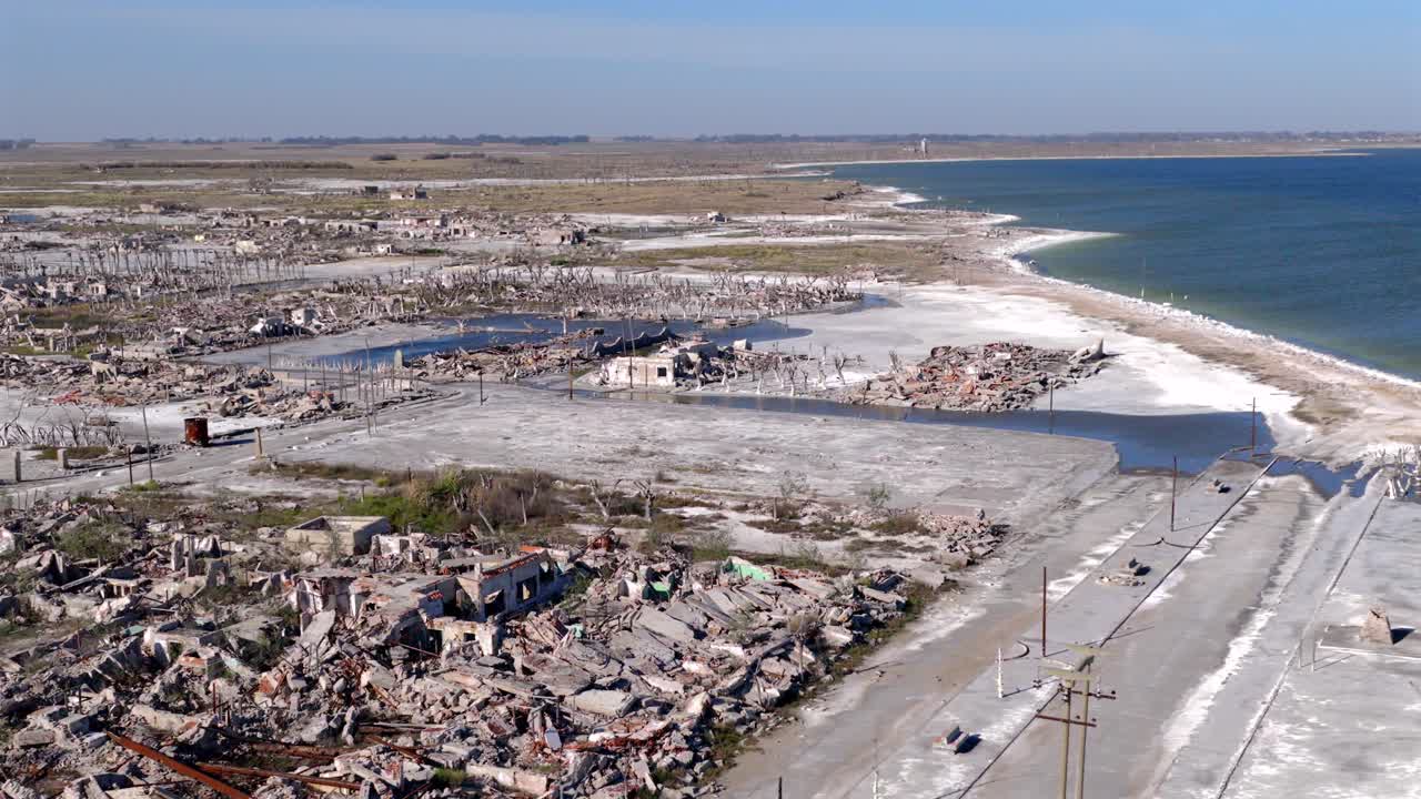 High wide drone shot spans abandoned ghost town of Villa Epecuén, where salt residues mark grid of collapsed buildings and skeletal streets, extending to the shoreline of Lake Epecuén, Argentina