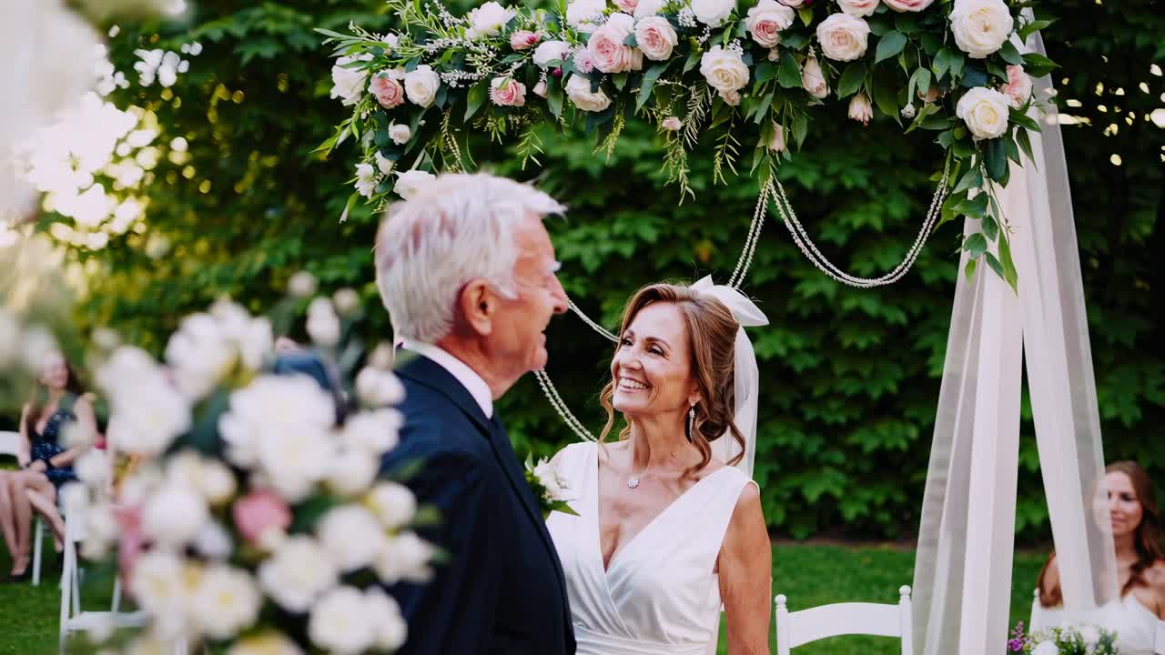A romantic wedding video scene captured at eye level, featuring a joyful couple under a floral arch