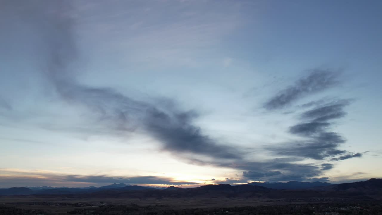 A sweeping landscape view featuring a mountainous horizon bathed in the warm glow of either sunrise or sunset. The sky is filled with layered clouds