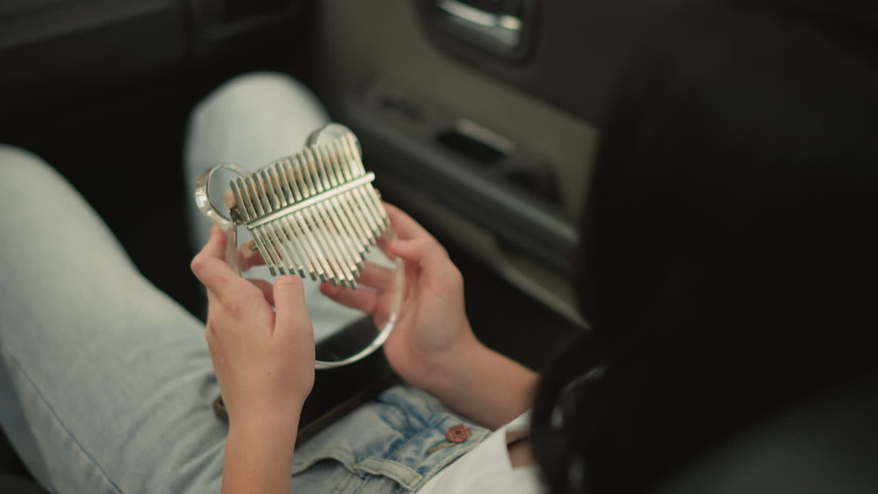 Unas manos sostienen una kalimba con forma de corazón sobre el regazo de un pasajero sentado en un coche. Las púas doradas atrapan la luz del sol, mientras que unos suaves rasgueos evocan un ambiente romántico. Viste un conjunto informal de jeans y el reflejo del instrumento añade brillo al viaje.
