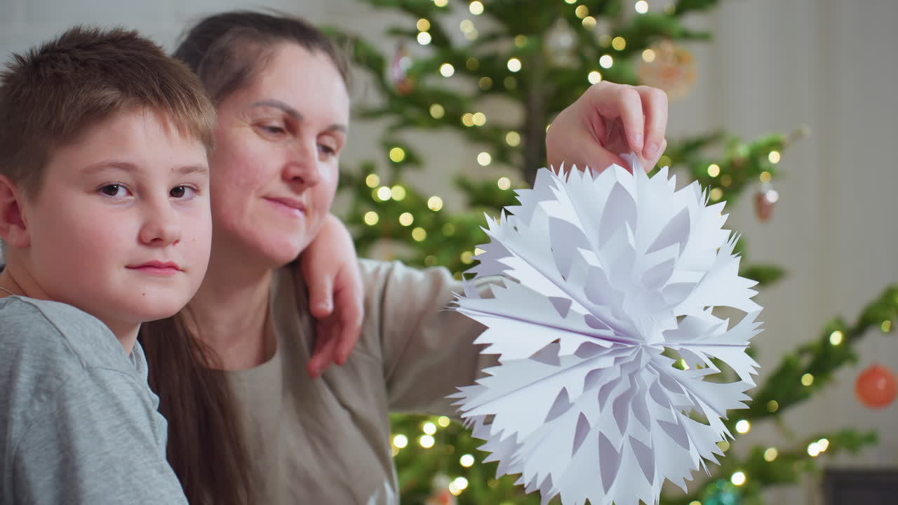 boy in grey top wraps arm around mother s shoulder while mother lifts crafted paper snowflake near decorated christmas tree both sharing calm moment indoors during festive holiday season