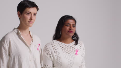 Studio-Portrait-Of-Two-Women-Wearing-Pink-Breast-Cancer-Awareness-Ribbons-Against-White-Background