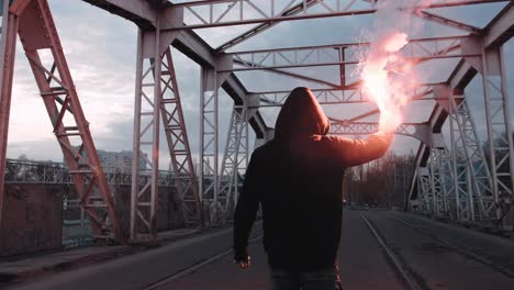 young man in hoodie and balaclava with red burning signal flare on the road under an old steel frame bridge, slow motion