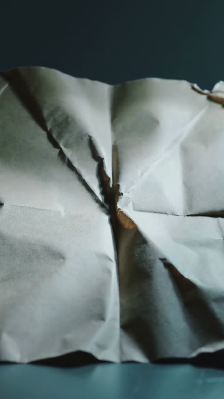 Vertical video: Shifting camera capturing creased white paper on blue table, revealing charred tear