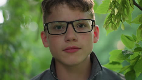 primer plano de un joven guapo con labios rosados, con gafas y una camiseta gris, mirando enfocado directamente a la cámara, está rodeado de vegetación con árboles borrosos en el fondo