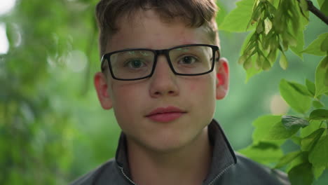 close-up of handsome young boy with pink lips, wearing glasses and a grey top, looking focused directly at the camera, he is surrounded by greenery with blurred trees in the background