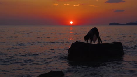 Junge-Frau,-Die-Yoga-Übungen-Auf-Einer-Großen-Klippe-Macht,-Umgeben-Von-Meerwasser,-Das-Den-Roten-Himmel-Des-Sonnenuntergangs-Widerspiegelt