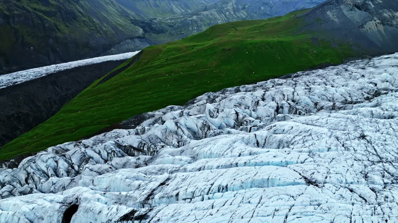 Luftdrone over en robust islandsk isbre, som avslører dype sprekker og blå is ved siden av levende grønne fjellsider og en vulkansk dal