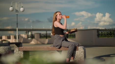 mujer sentada en un banco bebiendo de una botella de jugo mientras continúa leyendo un libro, la escena presenta un puente superior y un fondo borroso con postes de lámpara