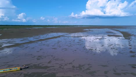 Aerial-fly-over-Tonle-Sap-Lake-during-low-water-levels-with-fishing-boat-moving-out-of-scene,-blue-sky-with-fluffy-clouds-on-horizon