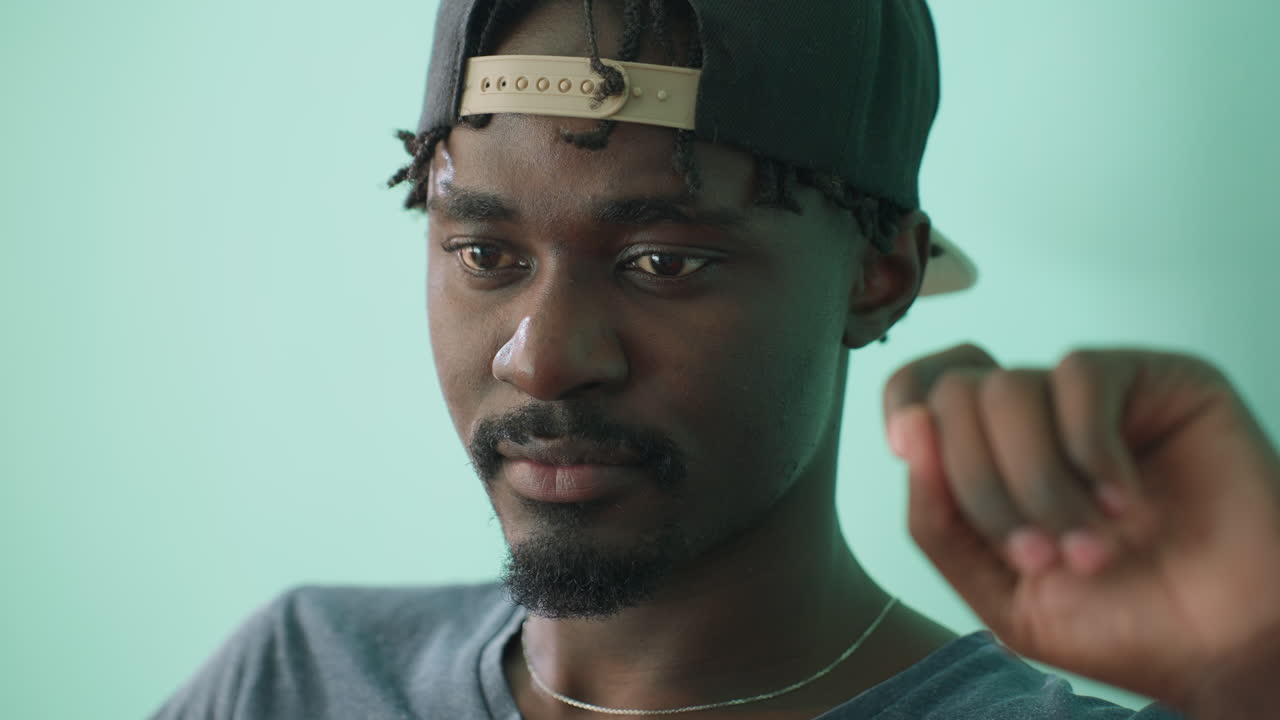 Close up of young man wearing cap and casual grey shirt sitting indoors against teal wall, lost in thought with pensive gaze, hand raised near face as he contemplates quietly